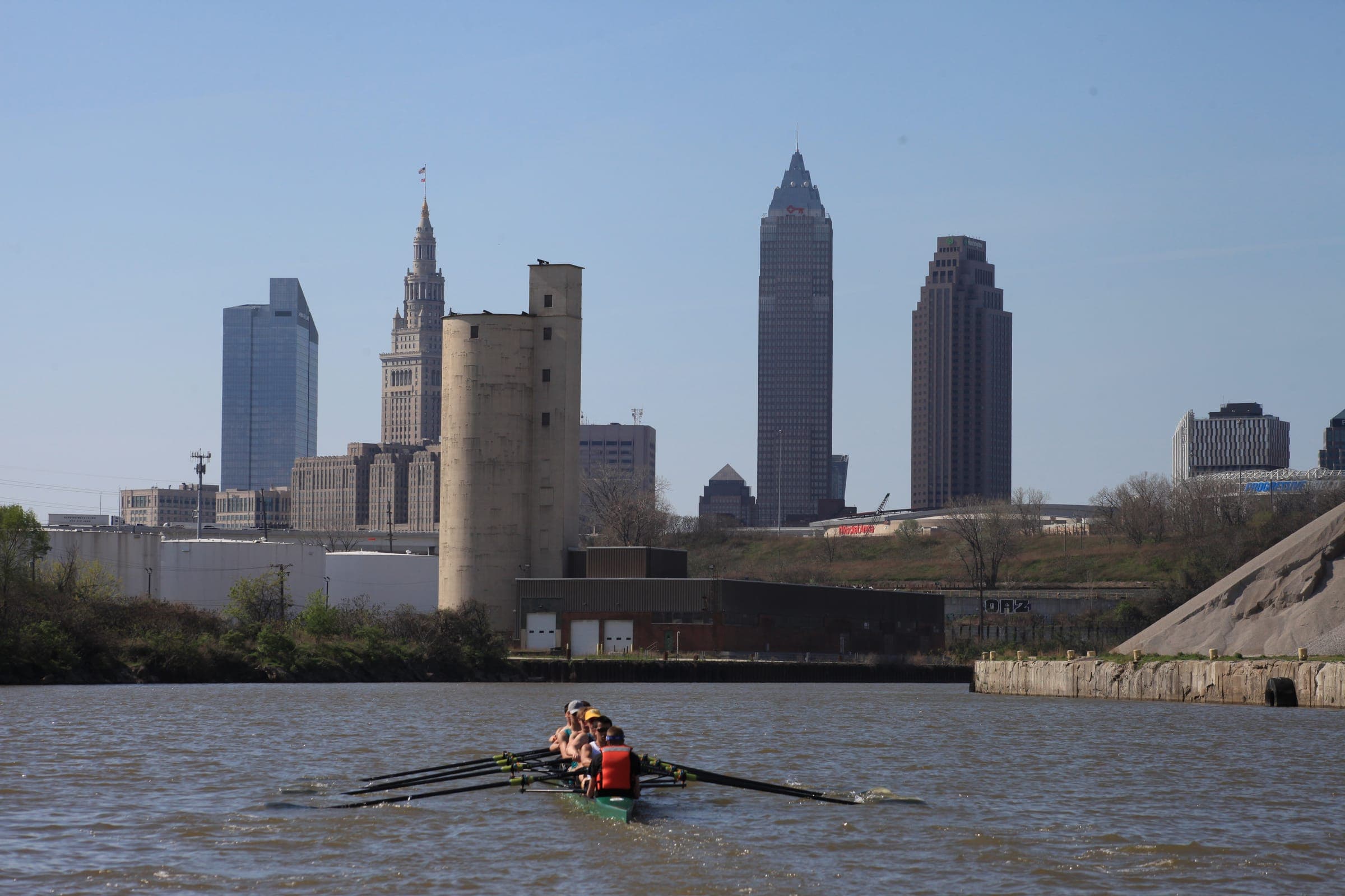 A crew rowing a boat down a river, with a city skyline behind them.